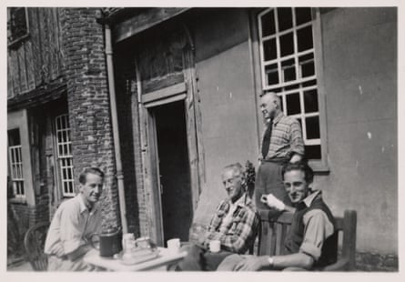 Black and white photo of four men of varying ages outside a farmhouse-style building. Three sit around a table with a teapot and mugs while a fourth leans against a wall. They all have their shirtsleeves rolled up and look relaxed in the sunshine.