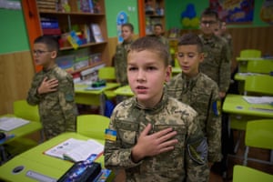 Young boys in military uniform stand beside their desks in a classroom and sing with their hands on their hearts