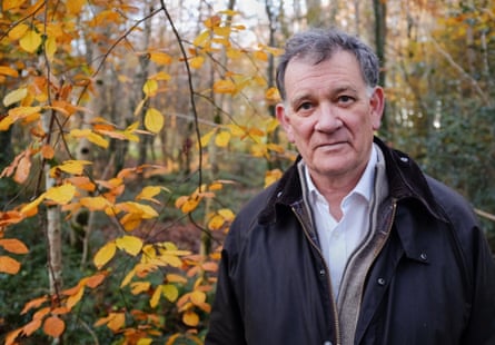 Man with shirt and dark overcoat standing among autumnal trees.