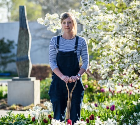 A woman in blue dungarees standing in a plant bed with a gardening fork, a sculpture behind her