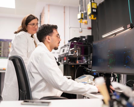 Seated man in white lab coat works at a monitor, as standing woman in white coat looks on.