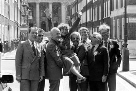 A black and white photograph of Shirley Williams being lifted up in the street by Steve Race and Barry Cryer, while Simon Cadell, Robert Powell, Denis Quilley, Richard Attenborough and Bamber Gascoigne stand next to them