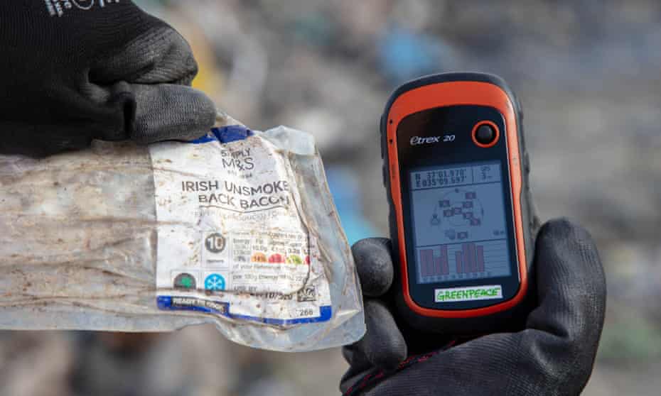 Hands holding up an empty back of M&S bacon and a device showing location