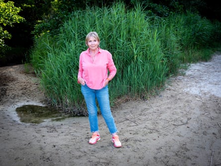 A woman wearing brightblue jeans, pink loose shirt and pink trainers standing in front of foliage with sand under feet