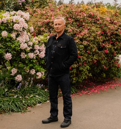 Robert Carlyle standing on a driveway, hands in jacket pockets, flowering bushes behind him
