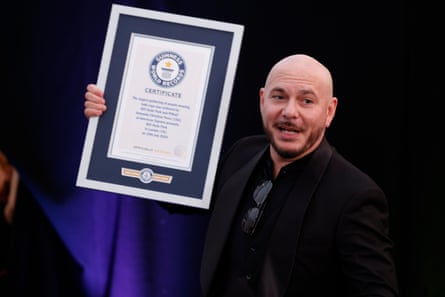 Pitbull, who is bald with a short dark beard, holds up his certificate. He wears a black suit and has sunglasses tucked into his collar.
