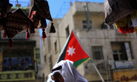 A man in a keffiyeh headdress walks in front of a Jordanian flag