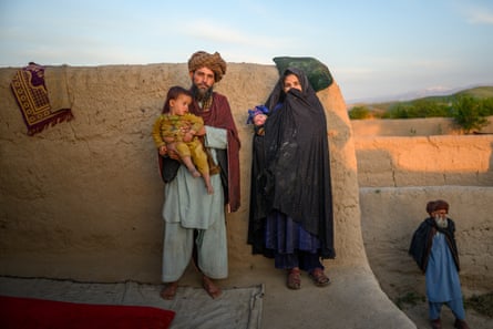 A girl holding a baby stands next to a man holding a toddler, outside a low mud house.