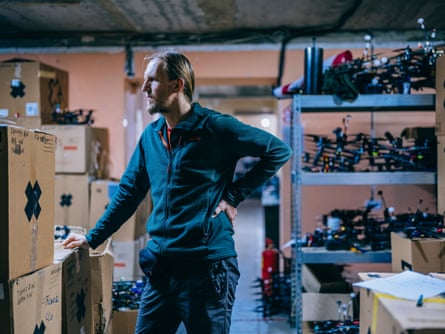 Dmytro, tall with a ponytail, in the store room of the workshop with lots of drones and boxes of equipment stacked up behind him
