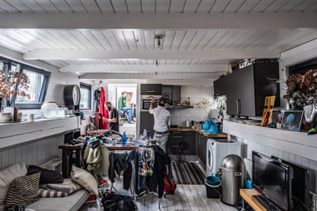 The three men are spread out in the messy houseboat with clothes strewn over chairs next to a dining table in the kitchen and cushions falling off the sofa in the foreground