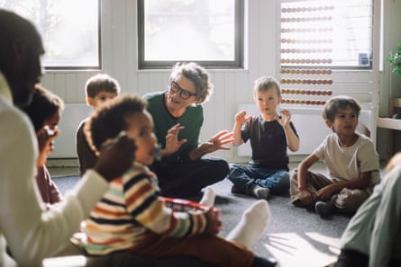 A senior female teacher playing with group of preschool children while sitting in classroom at kindergarten
