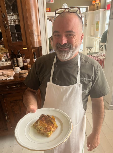 A smiling man in a white apron holds a plate of food.