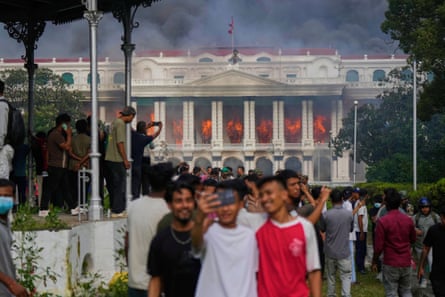 Young men celebrate and take photos in front of a white government building with a classical-style facade and pillars, which has flames and smoke pouring from it