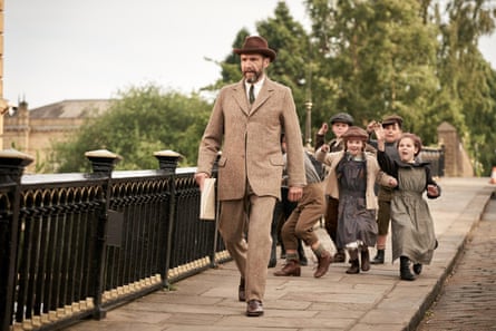 A film still of Ralph Fiennes looking troubled, dressed in a beige suit, white shirt, dark-green tie and brown hat, with some papers in his left hand, walking across a bridge in Yorkshire in 1916 with a group of young children running after him, jeering