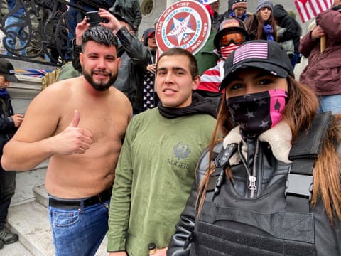 Two men and one woman in a row smiling for a selfie. Woman wears black hat with American flag, black and pink mask obscuring her face, black flak jacket and black leather jacket.
