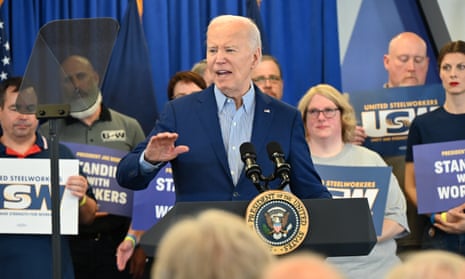 Older white man wearing blue suit, no tie, cuts air with hand as he speaks into mics at lectern with presidential seal, with people lined up and holding pro-union signs on stage behind him.