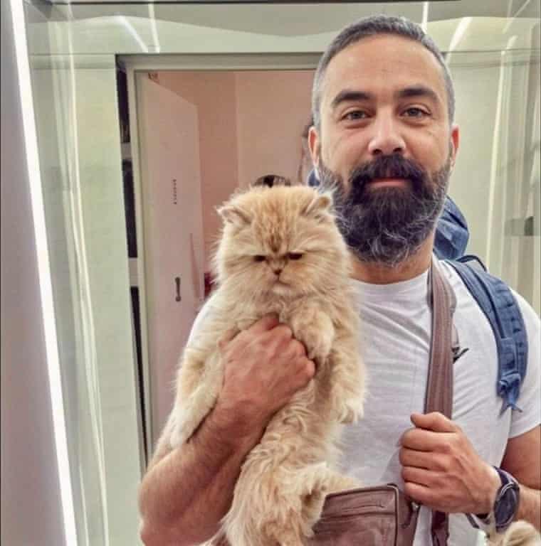 Ali Sina Mostafavi smiles while holding a fluffy cream-coloured cat indoors