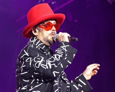 Boy George in a red hat and red sunglasses sings into a microphone wearing a black jacket with white graffiti-style text