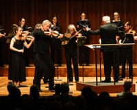 A conductor plays violin energetically while leading the Scottish Ensemble and singers on stage