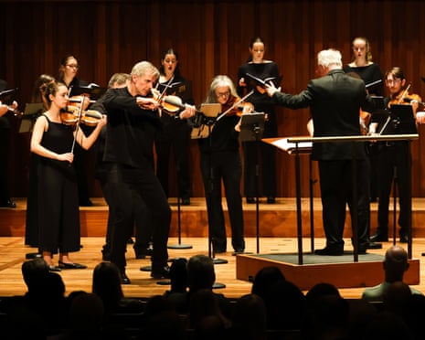 A conductor plays violin energetically while leading the Scottish Ensemble and singers on stage