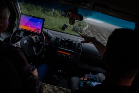 Someone points at a drone on the grass through a car windscreen with a computer set up on the dashboard