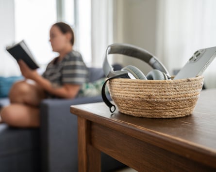 Basket with smartphone, smart watch and headphones on table with, in the background, a woman reading