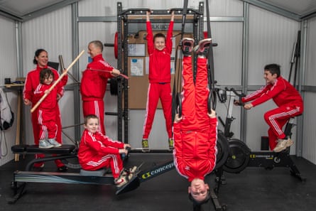 Two parents and five children, all wearing red tracksuits and using different equipment in a home gym: one on the rowing machine, one on the static bike, one hanging by his arms, another hanging upside down