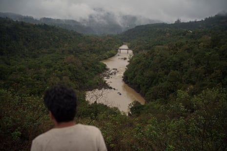 Rear view of a man’s head as he looks towards a river flowing through dense forest under cloudy skies.