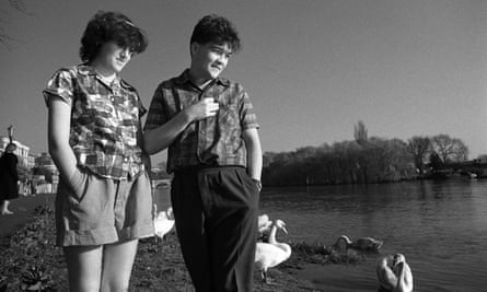 A black and white photograph of Tracey Thorn and Ben Watt by a lake in Richmond, with swans behind them. Tracey is wearing shorts and a short sleeved shirt. Ben is wearing trousers and a short sleeved shirt.