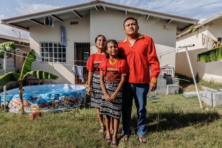 A man in a red shirt stands with a woman and a girl in traditional dresses on the grass outside a new-looking house
