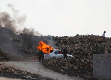 Israeli soldiers walk past a burning car