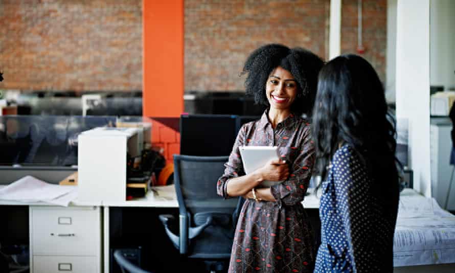 Smiling businesswoman standing at office workstation holding digital tablet in discussion with coworker