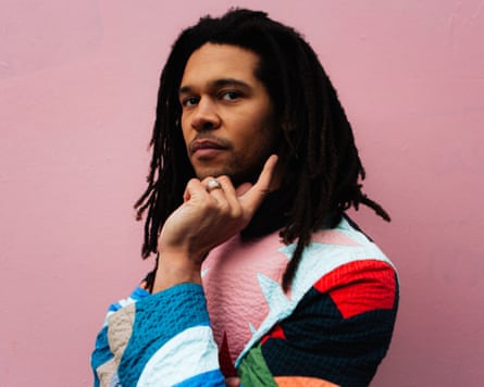 Standing against a pink wall, he holds his hand to chin and looks at the camera