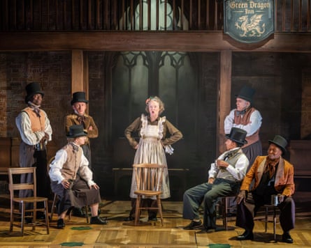 The cast of Middlemarch variously sit and stand on a stage while a woman in an apron stands front and centre looking angry.