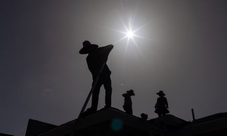 A deceptively black-and-white image, as the silhouettes of three people working on a roof, and the roof itself, are black in extreme shadow made by a blazing white sun.