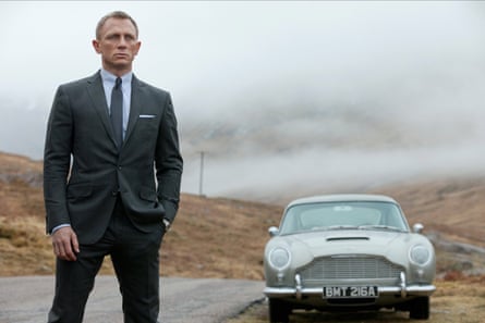 Daniel Craig as Jame Bond in Skyfall, 2012, standing by the side of a road with a sports car behind him