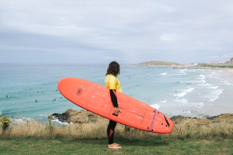Aiden Coxhead looks out over Fistral Beach, Newquay, with a red surfboard tucked under his arm.