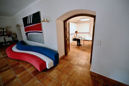he sits in the background at a table, framed by a doorway; next to the doorway is a modern sofa comprising three curved sections in stripes like the French flag, in red, white and dark blue.