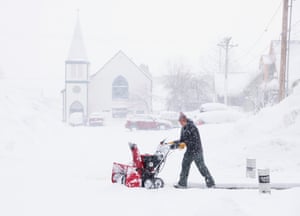 A man pushes a snow-clearing machine through snow. A church is visible through falling snow in the background