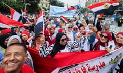 Women in a street in Giza, Egypt, many in sunglasses and all in headscarves, hold flags behind a banner, with a man grinning in front