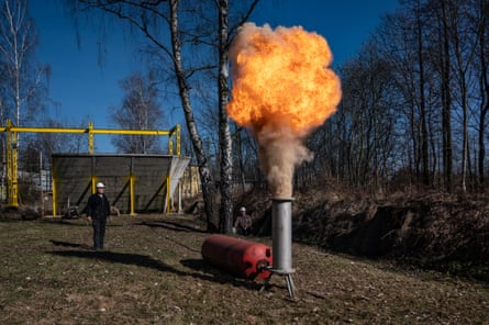 A large burst of flame erupts from a metal pipe in an outdoor, wooded area. Two people in protective gear stand nearby, observing the flare.