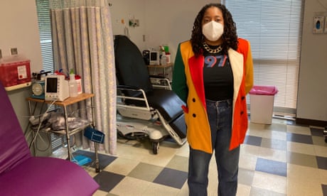 A woman in a colorful jacket, black T-shirt and jeans stands in a clinic with her hands behind her back
