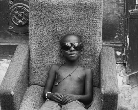 Mamadou, a shirtless boy in sunglasses, sits in his chosen armchair, hands folded