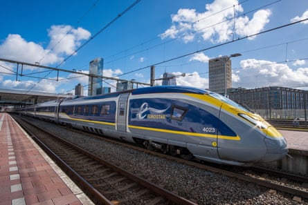 A blue, silver and yellow Eurostar train pulls up to a platform under a bright blue sky with small white clouds.