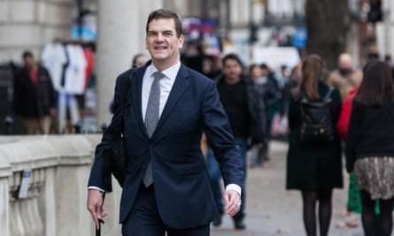 Oliver Robbins walking down a London street; he is wearing a dark suit and tie with white shirt; he is smiling and people are seen in the background. He is in his late 40s with short brown hair