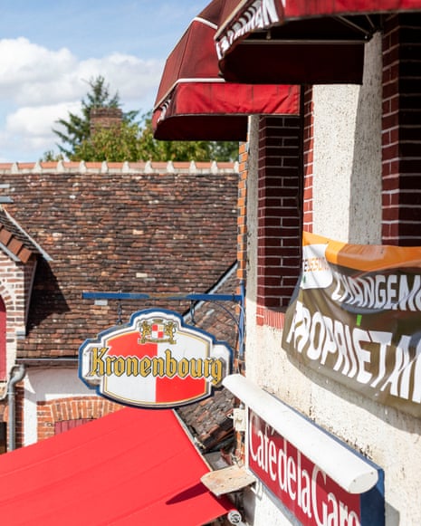A Kronenbourg sign hangs outside a French village café, with tiled roofs and red awnings beyond