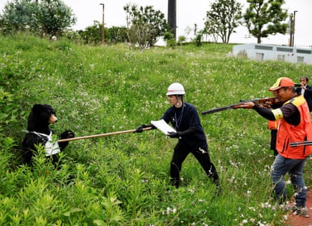 A man in a helmet uses a stick to prod a stuffed bear, while a hunter takes aim at the bear