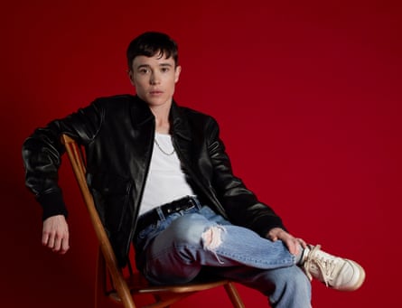 Elliot Page sitting on a chair wearing a leather jacket, jeans and a vest, against a red background