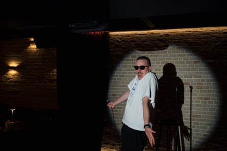 A man in sunglasses shrugs standing next to a mic stand.