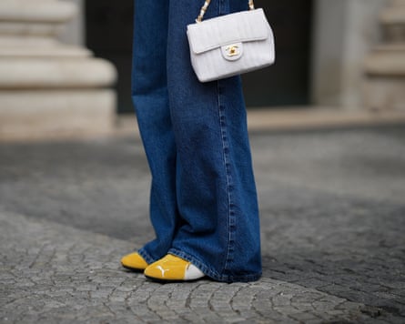 Pia Riegel is seen wearing a small white canvas, floral embroidered pattern bag with golden Chanel Logo and a white/golden carrier by Chanel, a dark blue long baggy denim pants with bronze button on the left trouser pocket by ZARA, yellow Puma Speedcat OG women sneakers
